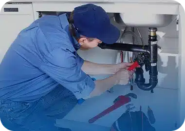 A plumber in a blue shirt and cap uses a wrench to work on pipes under a white kitchen sink.