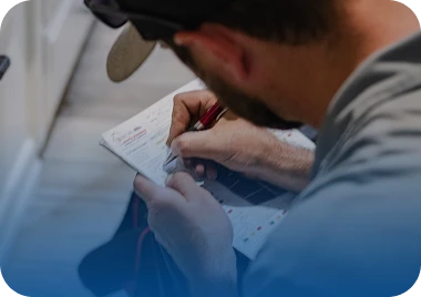 A man in a baseball cap writes on a white document with a red pen, focused on the task.