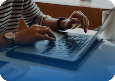A person wearing a striped shirt and watch types on a silver laptop with both hands.