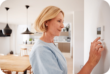 A smiling blonde woman in a light blue shirt adjusts a white digital thermostat on a white wall.