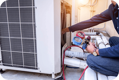 A technician in a dark blue shirt and jeans is working on an outdoor AC unit, holding gauges.