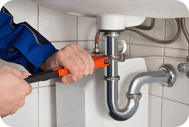 A person in a blue uniform uses an orange wrench to tighten a chrome pipe under a white sink.