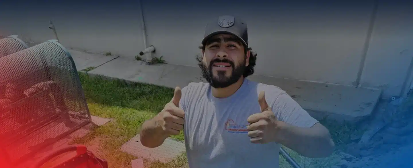 A smiling man with a beard and baseball cap gives two thumbs up, standing outdoors near a building.