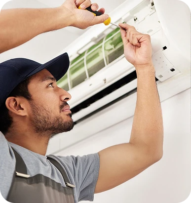 A man in a cap and overalls looks up as another person uses a screwdriver to repair an air conditioner.