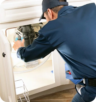 A plumber in a blue uniform and cap works on a garbage disposal unit under a kitchen sink.