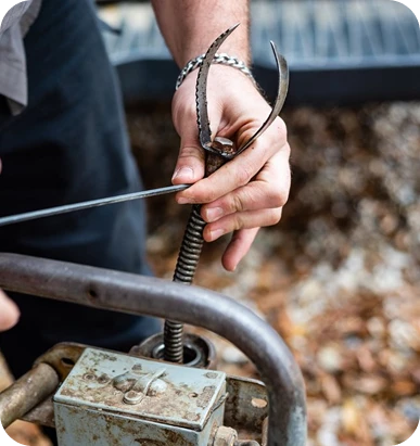 A person's hands are shown attaching a sharp, claw-like drain auger head to a coiled metal cable.