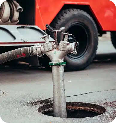 A metal fire hydrant with "2" on it and a hose connected, emerging from a manhole, with a red truck in the background.