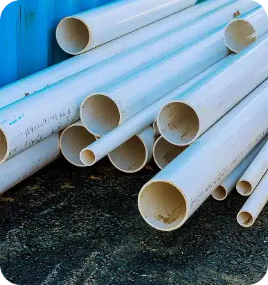 A pile of white PVC pipes of various diameters rests on dark asphalt, with a blue structure in the background.