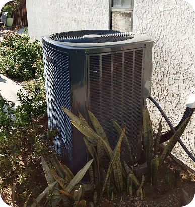 A gray outdoor air conditioning unit sits next to a white stucco wall, surrounded by green plants.