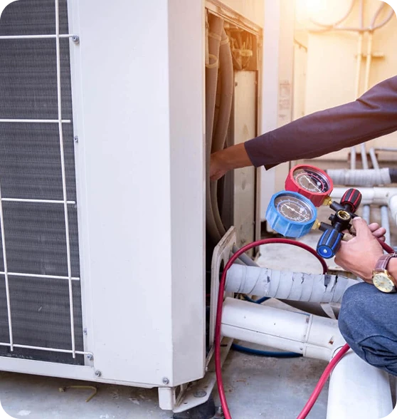 A technician in a dark shirt and jeans works on an outdoor air conditioning unit, holding a gauge manifold.