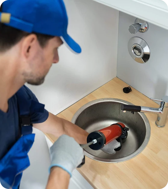 A plumber in a blue cap and overalls uses a red drain blaster to clear a stainless steel sink.