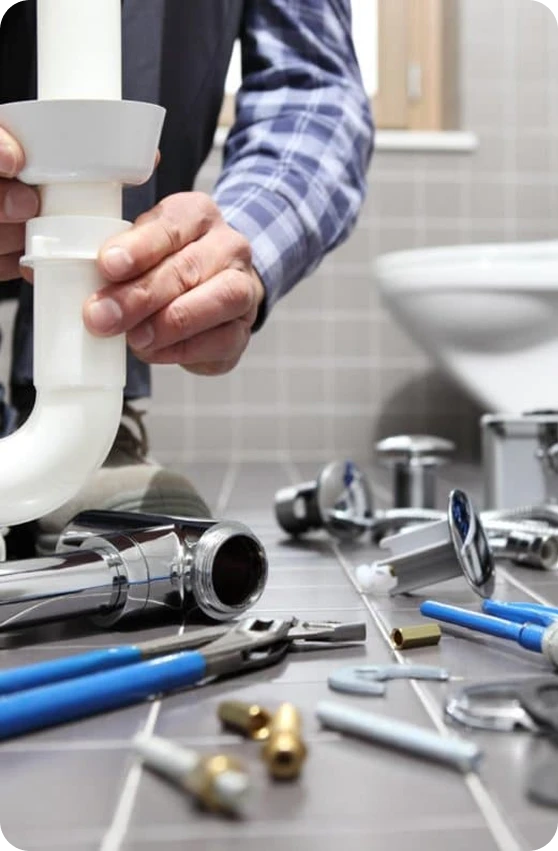 A plumber in a blue plaid shirt installs a white P-trap pipe, surrounded by various tools and chrome fixtures.