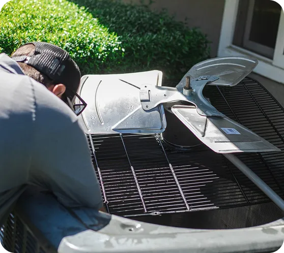 A person in a black cap and gray shirt leans over an AC unit with its fan blades removed.