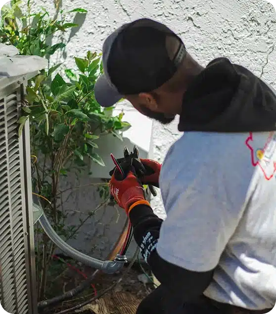 A person in a gray cap and orange gloves uses pliers to work on electrical wires near an AC unit.