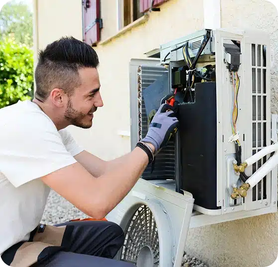 A smiling man in a white shirt and gloves works on an open outdoor air conditioning unit.