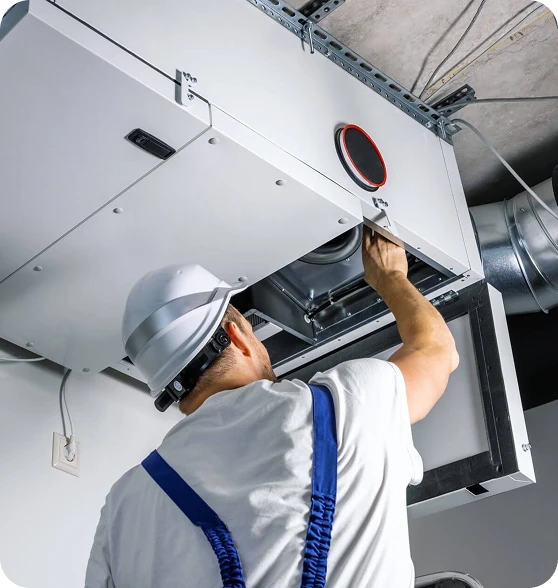 A man in a white hard hat and blue overalls works on a large white HVAC unit mounted on a ceiling.