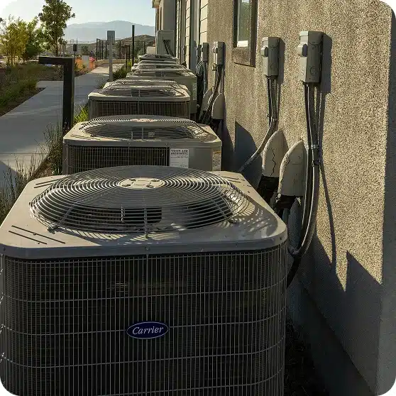 A row of "Carrier" air conditioning units with fans on top sit beside a building with electrical conduits.