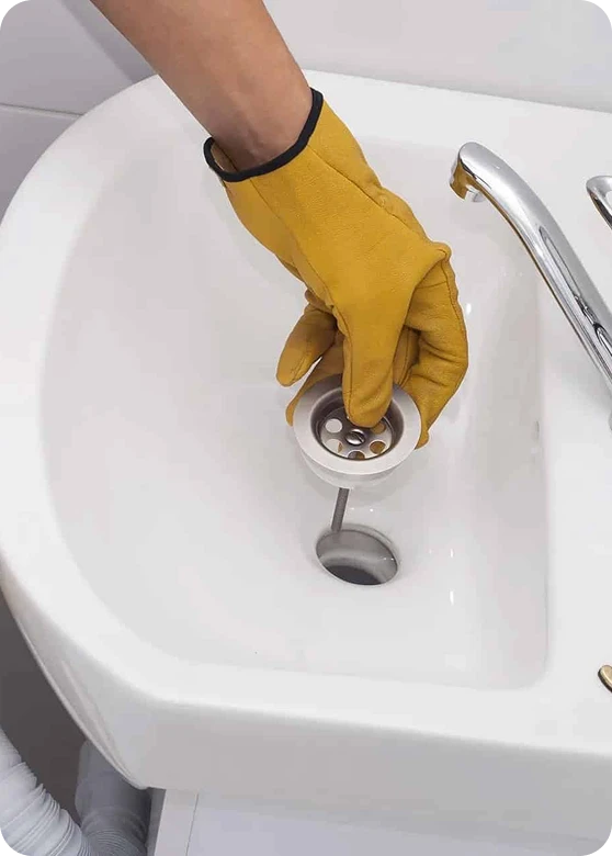 A gloved hand removes a metal drain stopper from a white bathroom sink with a chrome faucet.