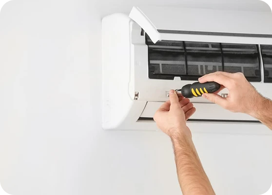 A person's hands use a screwdriver to remove a screw from an open air conditioner unit on a white wall.