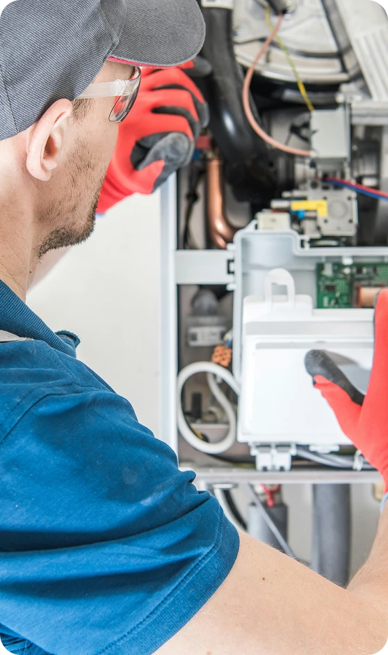 A man in a gray cap, safety glasses, and red gloves works on the exposed internal components of a boiler.