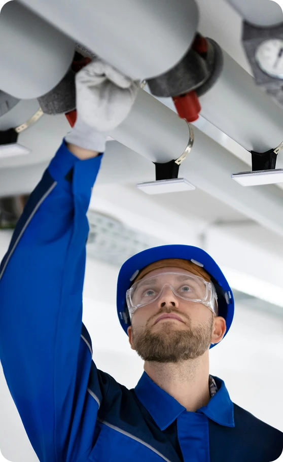A man in a blue hard hat, safety glasses, and work uniform reaches up to adjust ceiling pipes.
