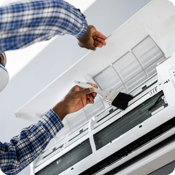 A person in a plaid shirt cleans the inside of an open wall-mounted air conditioner with a brush.