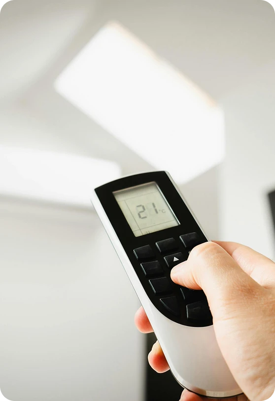 A hand holds a white and black remote control displaying "21°C" on its screen, with a bright white ceiling in the background.
