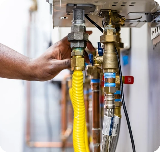A dark-skinned hand adjusts a brass fitting on a yellow corrugated gas line connected to a water heater.