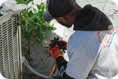 A man in a gray cap and red gloves uses pliers to work on wires next to an AC unit.
