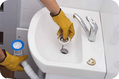 A person in yellow gloves installs a sink drain, holding the top part over the basin and the bottom part below.