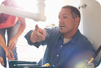A man in a blue shirt points while looking at a white pipe under a sink, with a woman nearby.