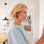 A smiling blonde woman in a light blue shirt adjusts a white digital thermostat on a white wall.