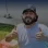 A smiling man with a beard and baseball cap gives two thumbs up, standing outdoors near a building.